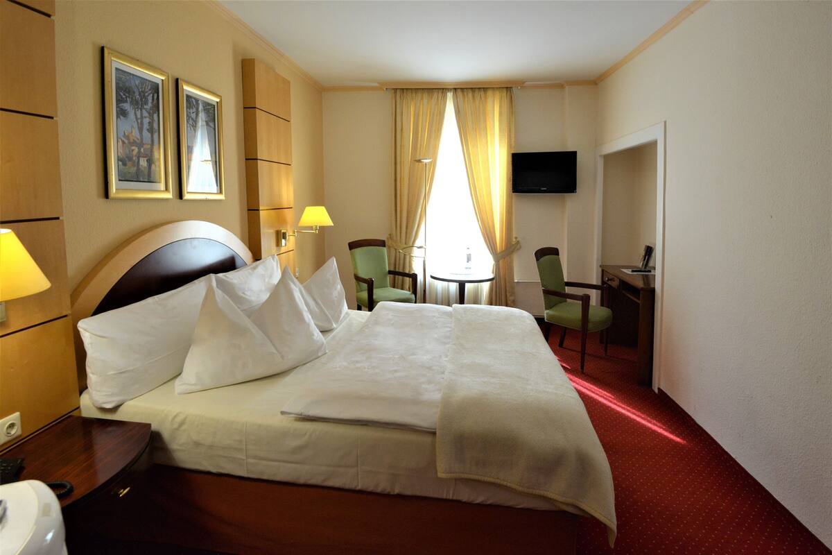 Une chambre d’hôtel avec un lit, deux chaises, un bureau, une télévision et deux tableaux encadrés au mur.