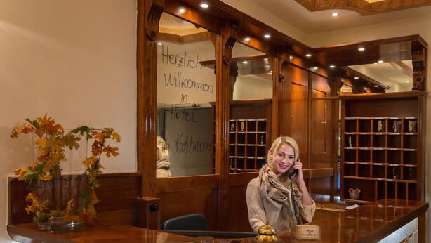 Een receptiebalie met houten afwerking, een vaas met herfstbladeren, en een bord met de tekst 'Herzlich Willkommen in Hotel Kochbamber'.