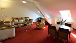 Een loft-woonkamer met een rode vloerbedekking, een zithoek, een salontafel, een eettafel met stoelen, en een groot raam dat natuurlijk licht binnenlaat.