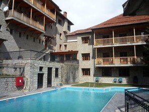 A multi-story building with a stone facade, balconies with wooden railings, and a swimming pool in the foreground.