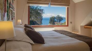 A bedroom with a large window, a bed with pillows, a bedside lamp, and a view of the sea.