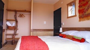 A bedroom with a bed, a wooden shelf with towels, a red blanket, and a window with a view. - Casa Kalfu (Puerto Varas)