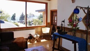 A cozy living room with a large window, a wooden table with a blue tablecloth, and a decorative wall piece. - Casa Kalfu (Puerto Varas)