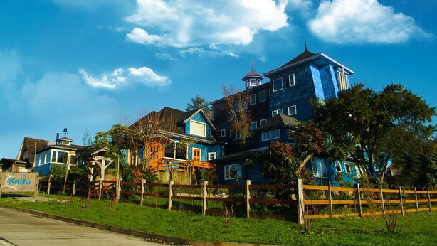A large, multi-story building with a distinctive blue facade, surrounded by a wooden fence and greenery. - Casa Kalfu (Puerto Varas)