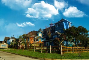 A large, multi-story building with a distinctive blue facade, surrounded by a wooden fence and greenery. - Casa Kalfu (Puerto Varas)