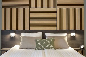 Una cama bien hecha con dos almohadas y un cojín decorativo, un teléfono en la mesita de noche y una lámpara fijada a la pared.