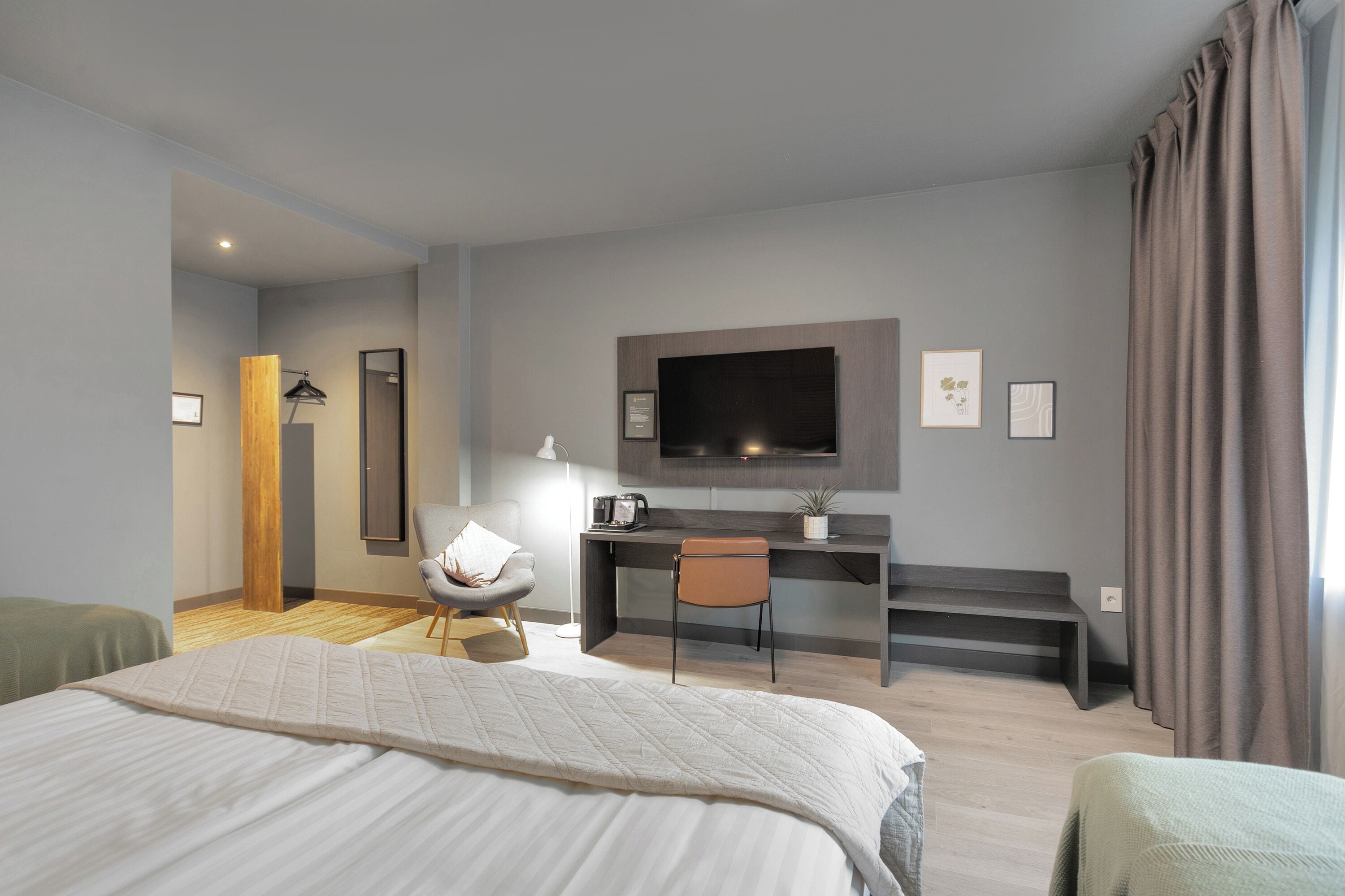 A modern hotel room with a large bed, a desk with a chair, a flat-screen TV mounted on the wall, and a window with curtains.