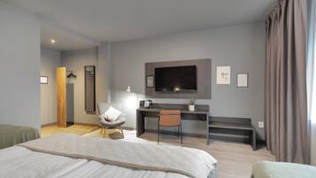 A modern hotel room with a large bed, a desk with a chair, a flat-screen TV mounted on the wall, and a window with curtains.