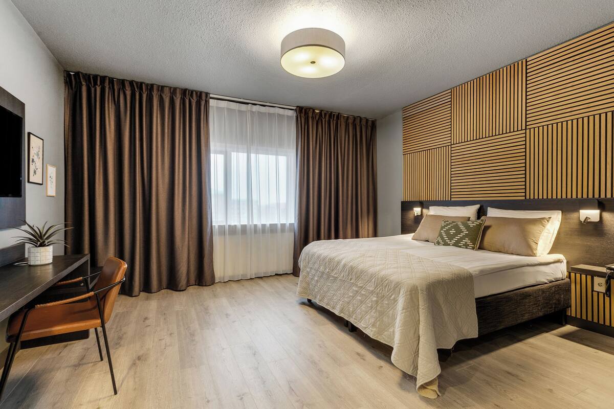 A modern hotel room with a wooden floor, a large bed with a headboard, a desk with a chair, and a large window with curtains.