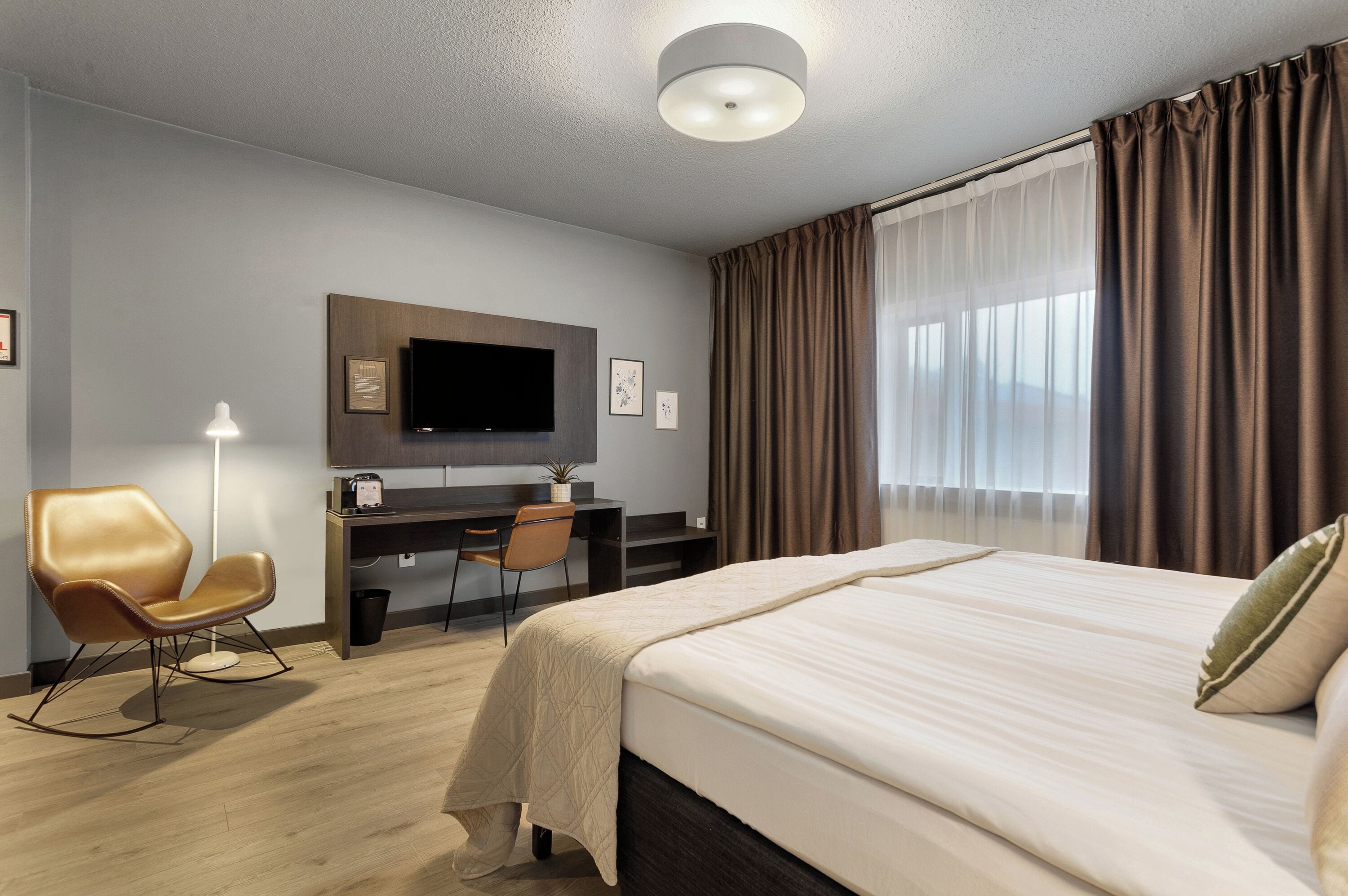 A hotel room with a large bed, a desk with a chair, a flat-screen TV mounted on the wall, and a window with curtains.