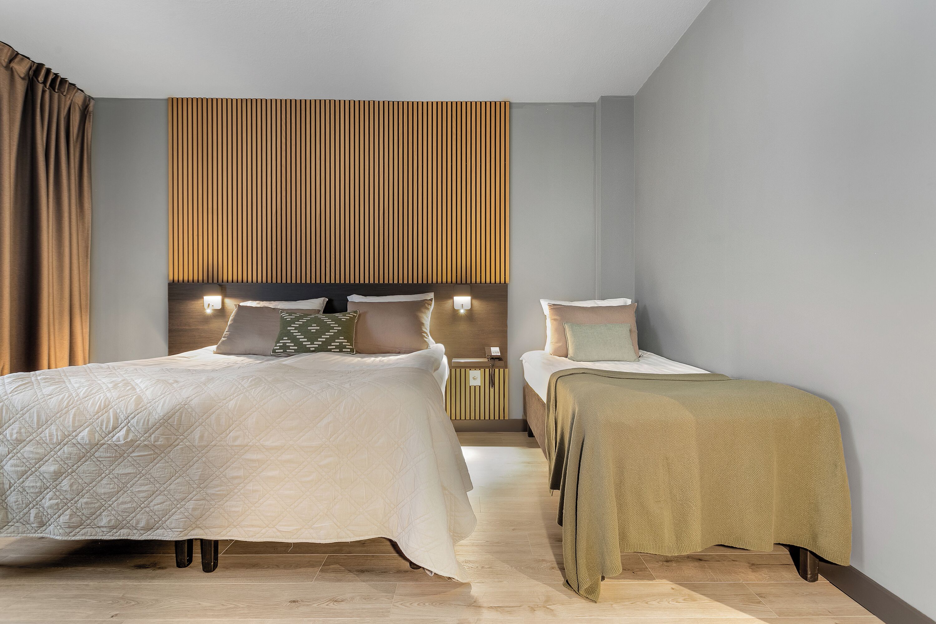 Ein Hotelzimmer mit einem großen Bett, einem kleineren Bett, einem Lattenrost und einem Nachttisch mit einer Lampe.