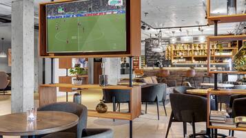 Zona de bar moderna amb un televisor gran que mostra un partit de futbol, mobles de fusta i prestatges amb ampolles.