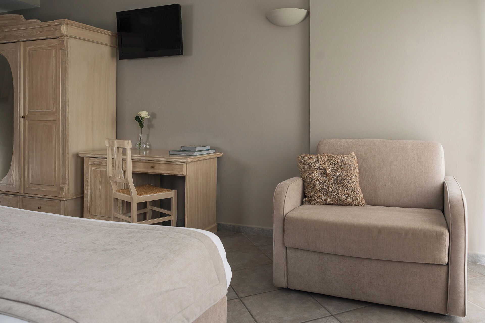 A hotel room with a bed, a desk with a chair, a sofa with a pillow, a television on the wall, and a vase with a flower on the desk.