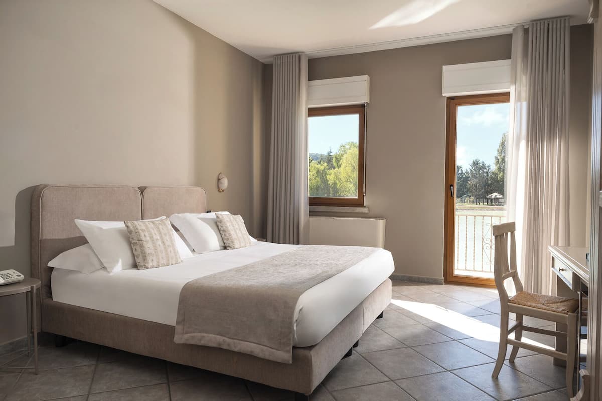 A hotel room with a large bed, a desk, and a view of a balcony with trees and a body of water.
