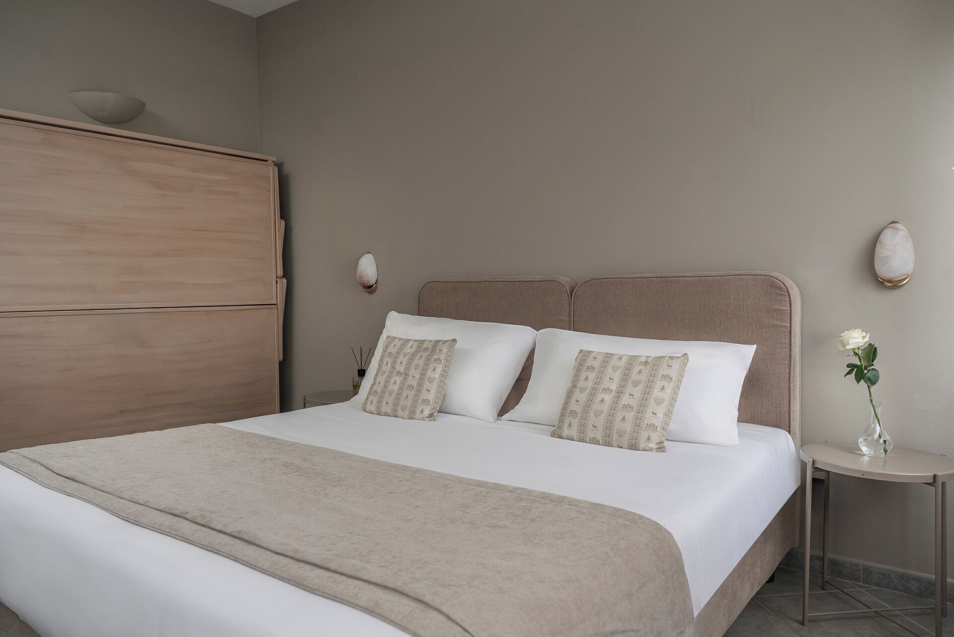 A neatly made bed with a beige blanket, two pillows with a patterned design, and a bedside table with a vase of flowers.
