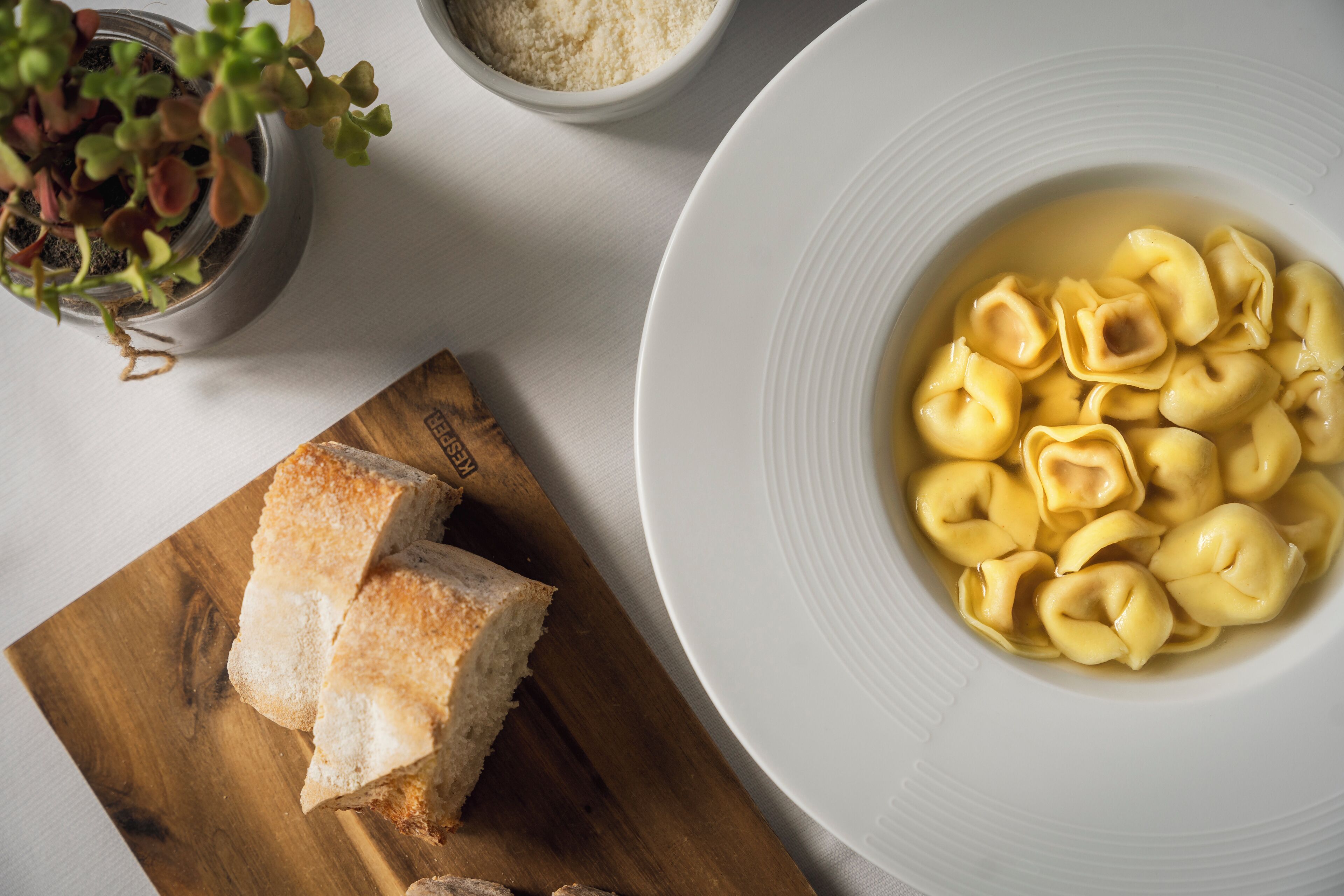 A bowl of tortellini in a white bowl, a wooden cutting board with bread slices, and a small potted plant with green leaves.