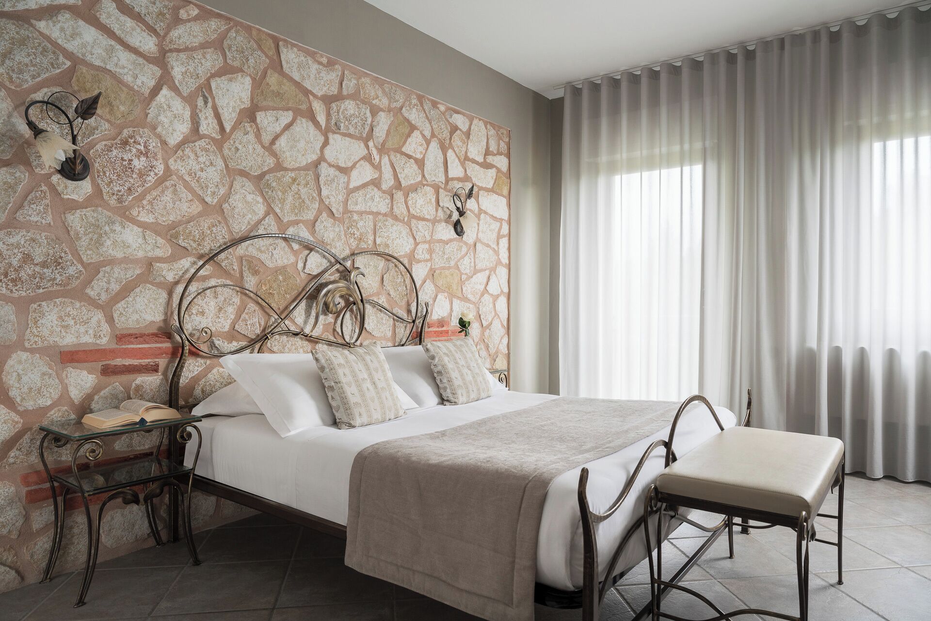 Un dormitorio con pared de piedra, cama con estructura de metal, mesita de noche y un banco.