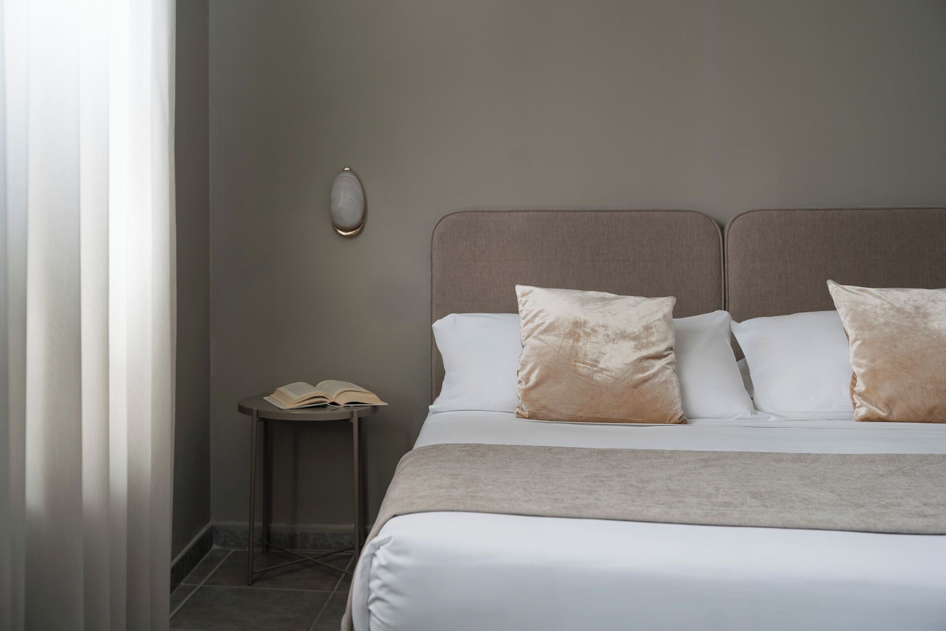 A neatly made bed with two pillows, a bedside table with an open book, and a wall-mounted light fixture.