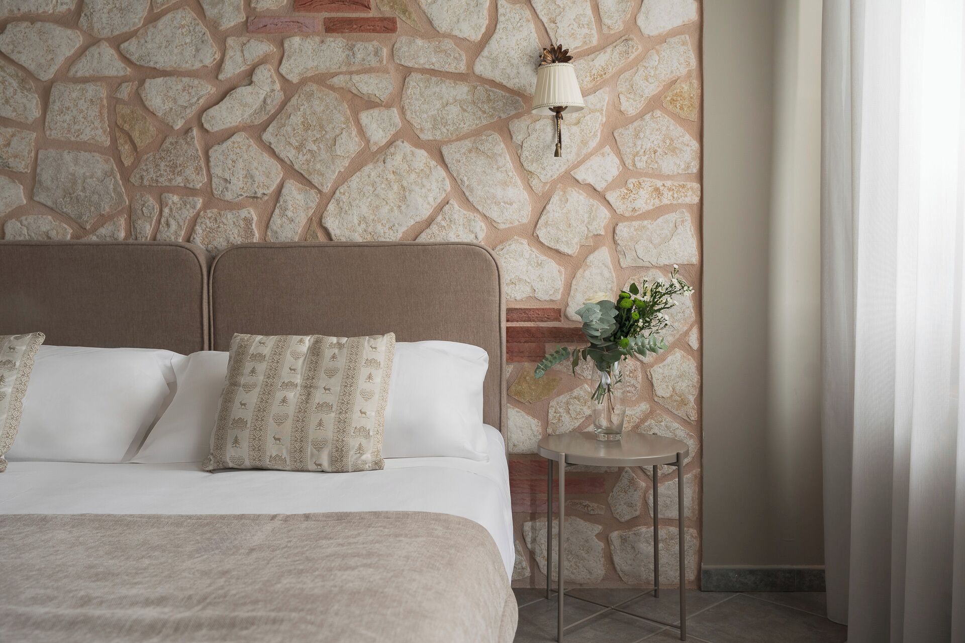 A bedroom with a stone wall, a bed with white bedding, and a small table with a vase of flowers.