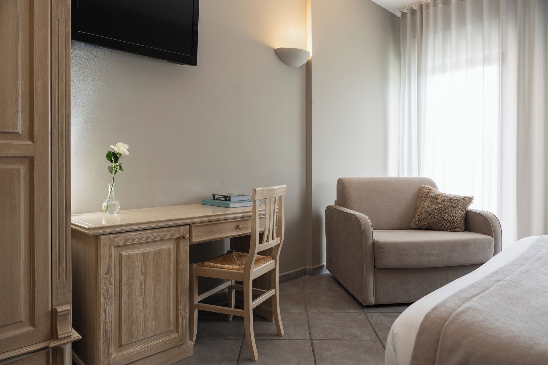 A hotel room with a bed, a sofa, a desk with a chair, a TV mounted on the wall, and a window with curtains.