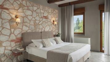 Un dormitorio con pared de piedra, una cama con ropa de cama blanca, dos lámparas de noche y una ventana con vistas a la vegetación.