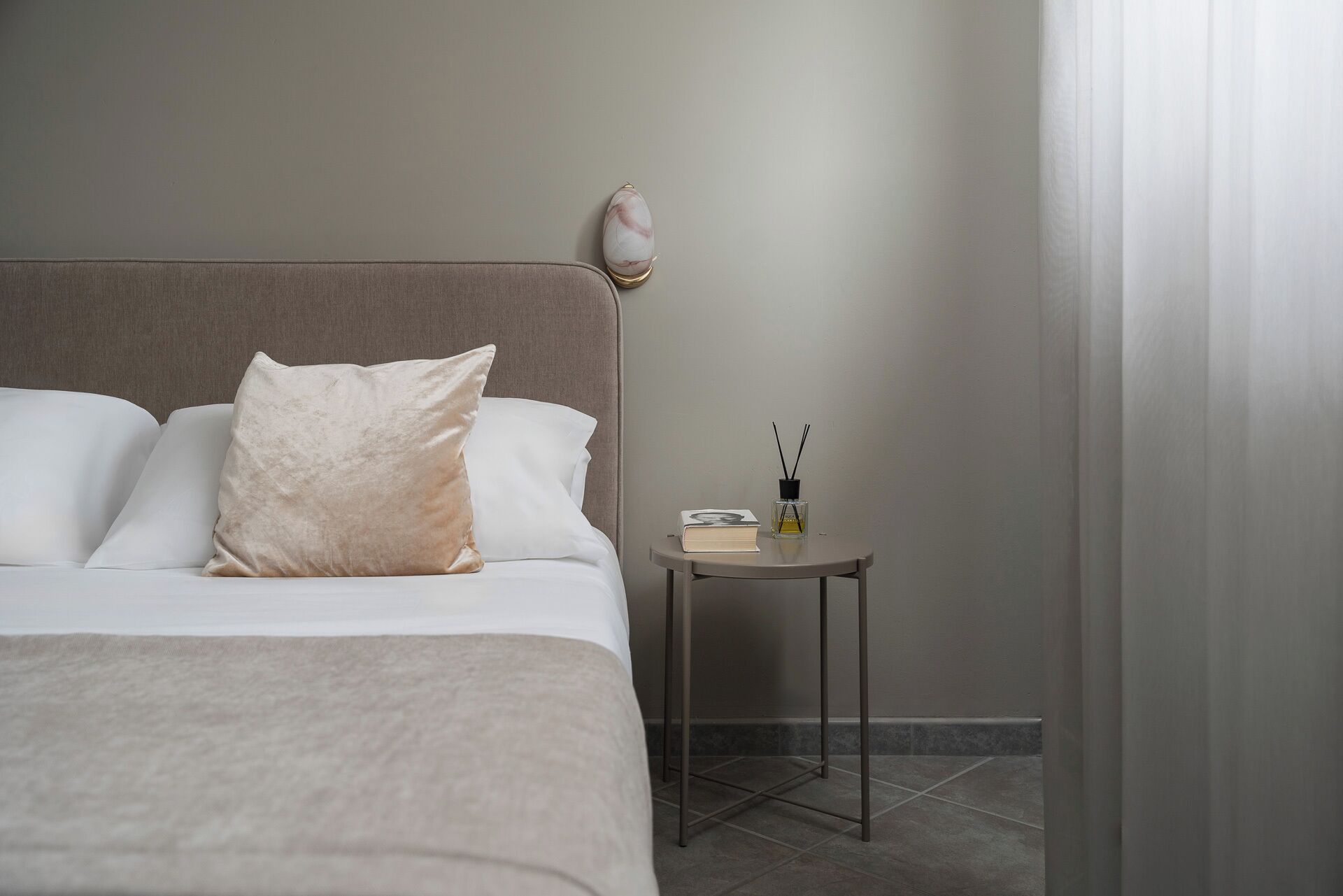 A neatly made bed with a large beige pillow, a small bedside table with a book and a bottle of perfume, and a plain wall with a curtain partially drawn.