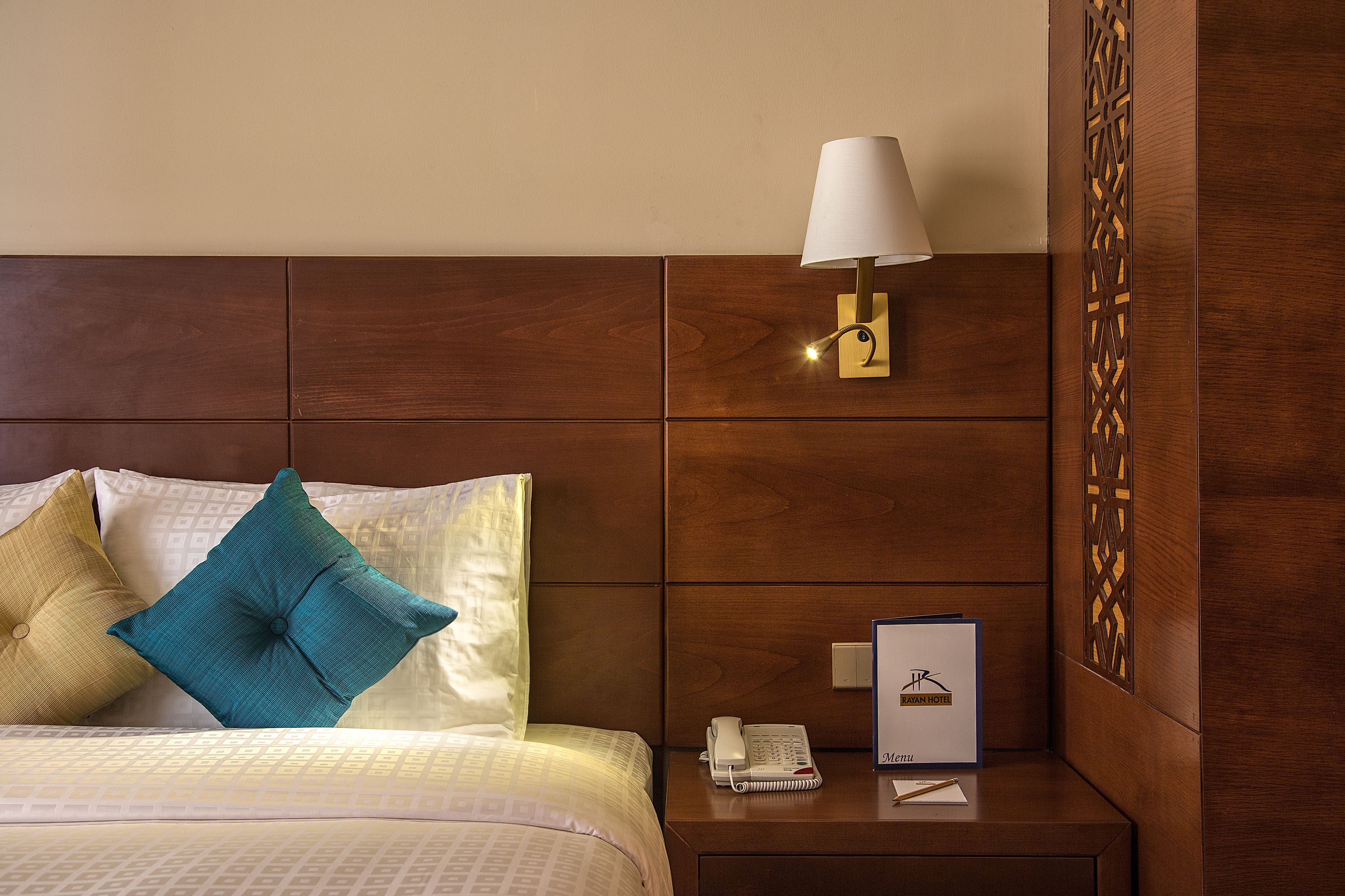 A neatly made bed with two pillows, a bedside table with a lamp, a phone, and a framed item.