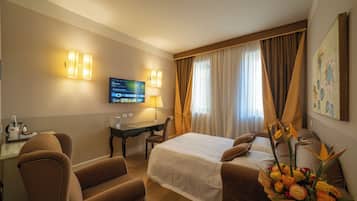 A hotel room with a bed, a desk, a chair, a TV, and a floral arrangement.