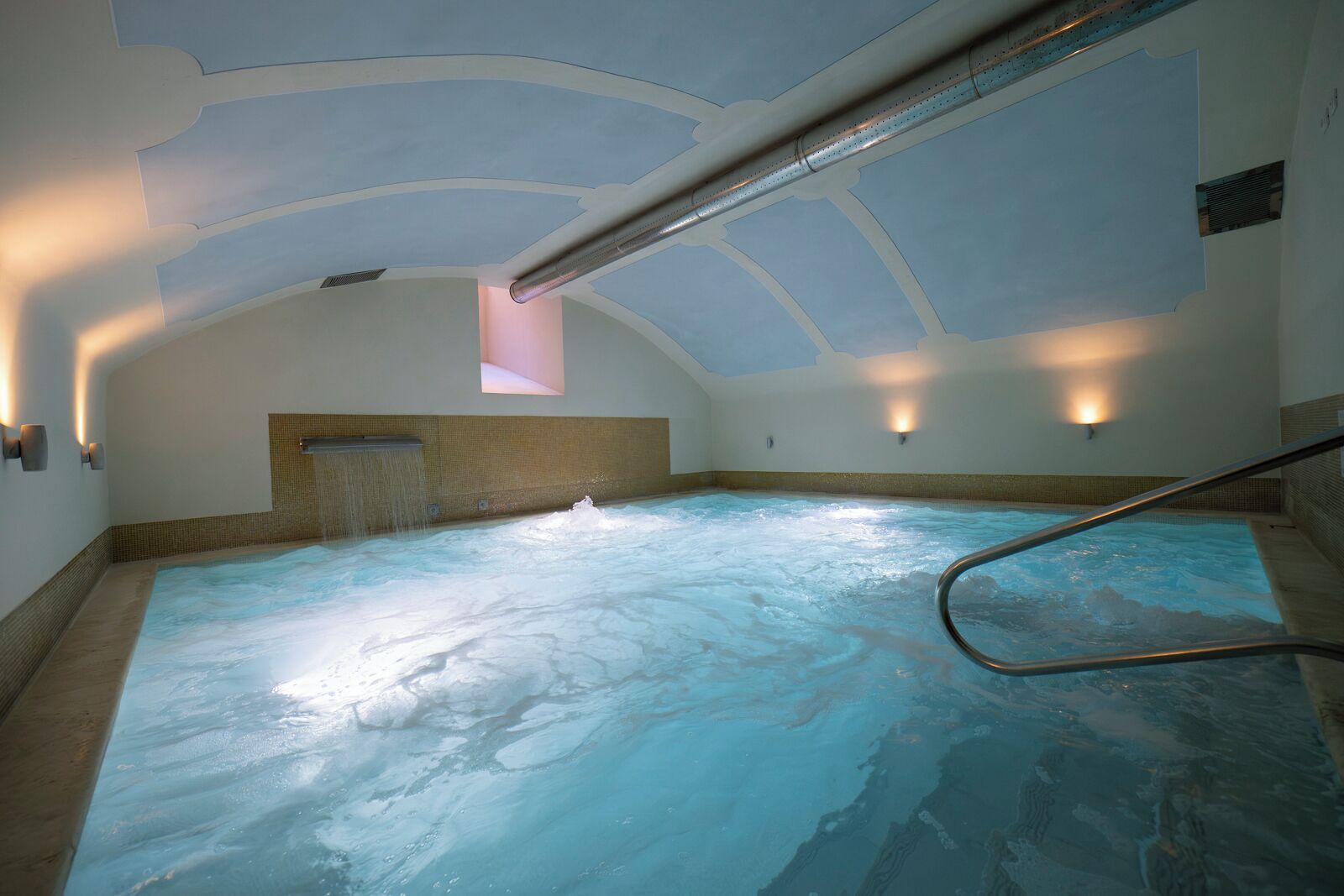 A hot tub with a waterfall feature, a metal railing, and a ceiling with a unique design.