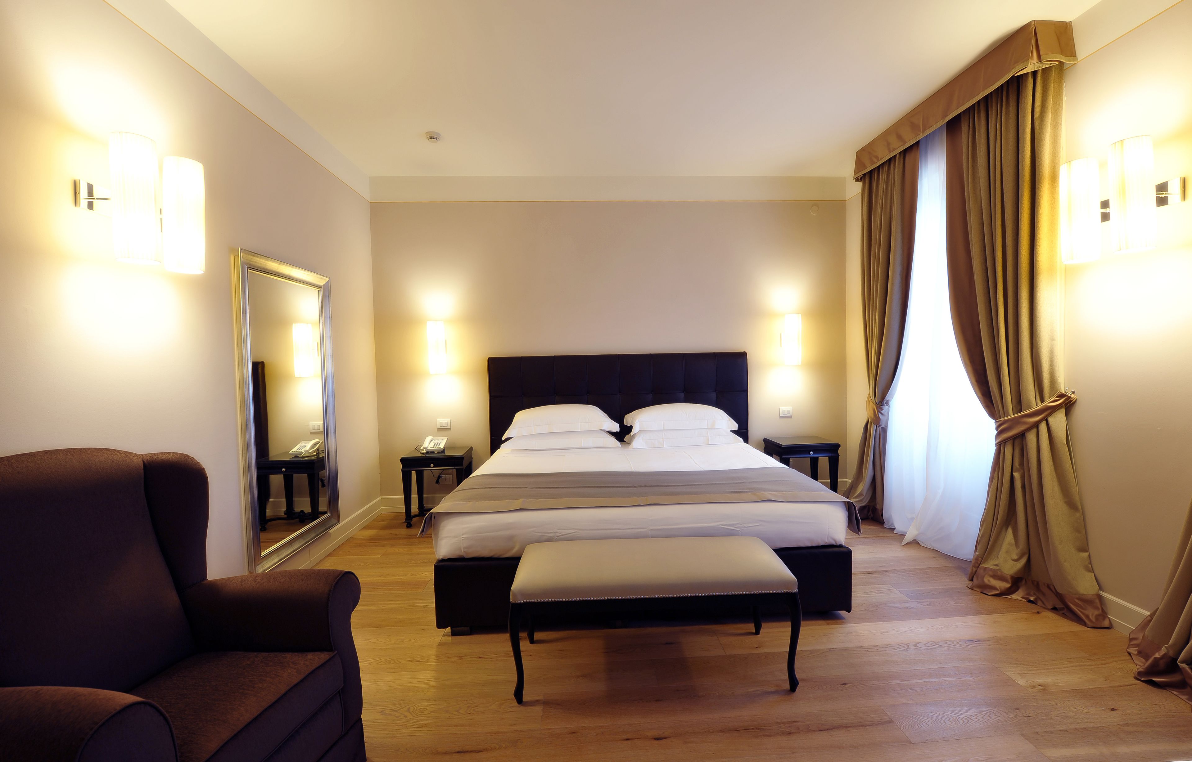 A hotel room with a large bed, a chair, a bench, and two bedside tables with lamps.