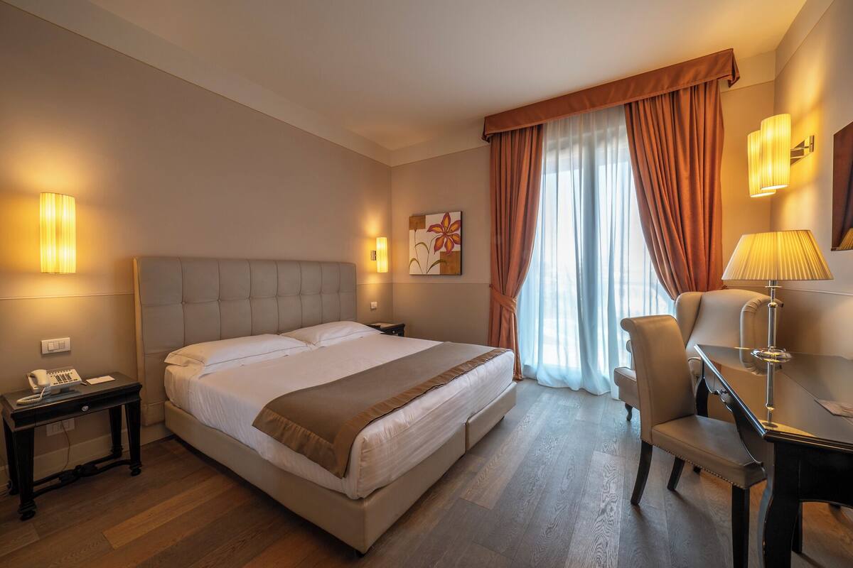 A hotel room with a large bed, a desk with a chair, a lamp, and a painting on the wall.