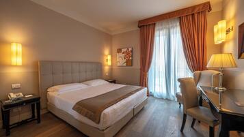 Una camera d'albergo con un letto grande, una scrivania con una sedia, una lampada e un quadro appeso al muro.