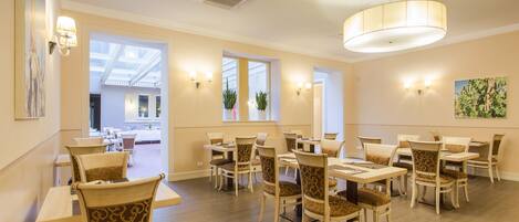 A dining area with multiple tables and chairs, large windows, and wall-mounted lighting.