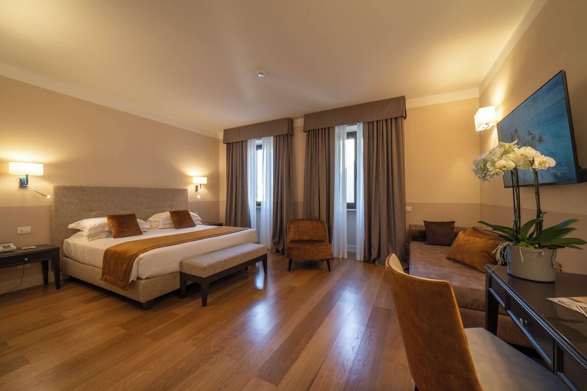 A hotel room with a large bed, a chair, a desk, and a potted plant.