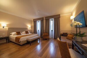 A hotel room with a large bed, a chair, a desk, and a potted plant.