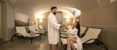 A spa room with reclining chairs, a man and a woman in robes, and wall-mounted lights.