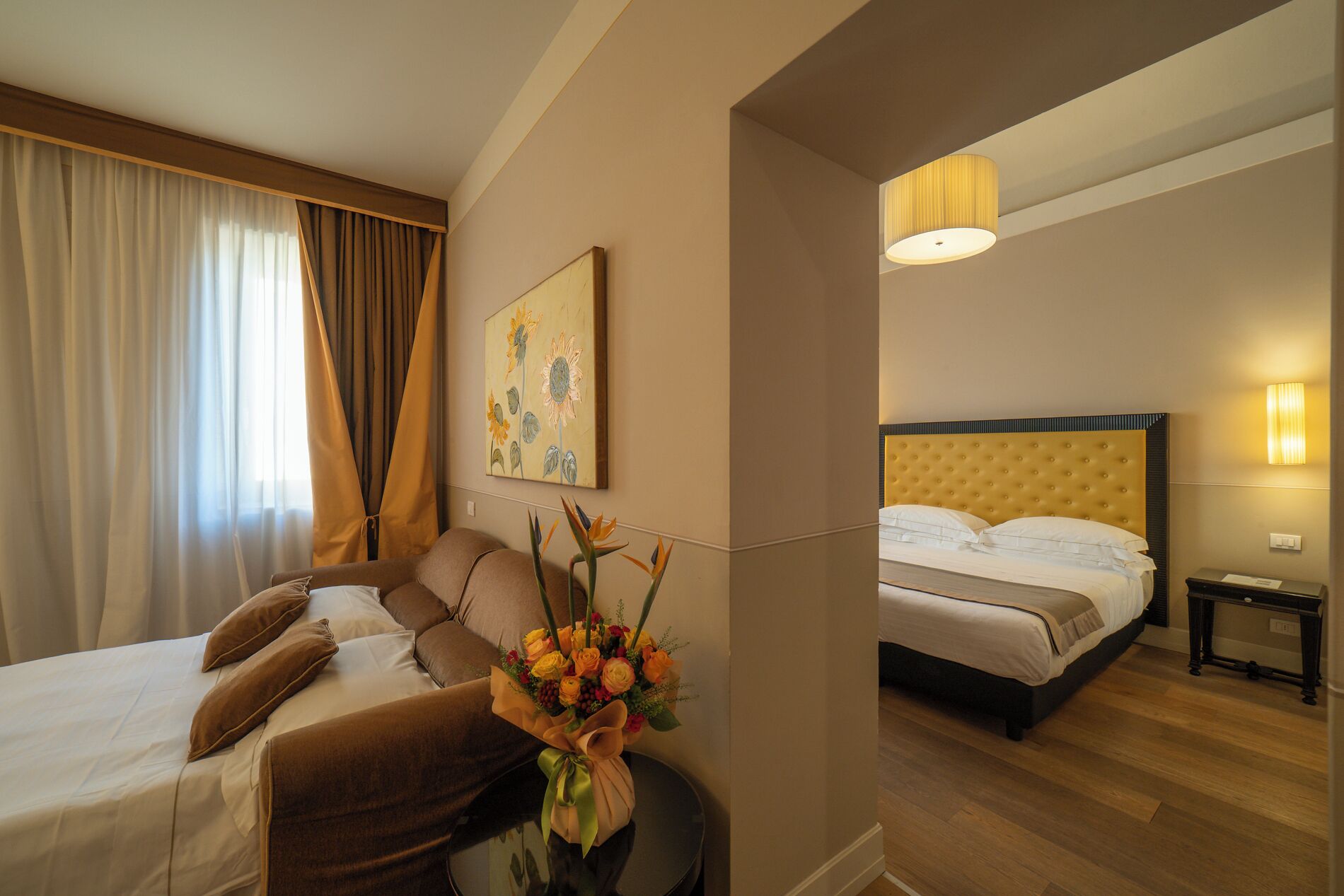 A hotel room with a bed, a sofa, a small table with a bouquet of flowers, and a painting on the wall.