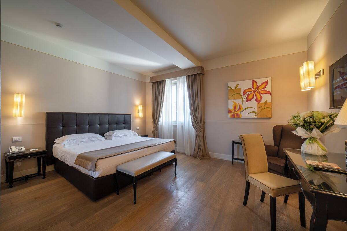 A hotel room with a large bed, a desk, a chair, and a painting on the wall.