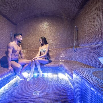 A couple seated in a tiled hot tub with a mesh cover and a showerhead visible.