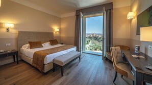 Habitación de hotel con una cama grande, un escritorio, una silla y vista a la ciudad por la ventana, además de una lámpara en el techo.