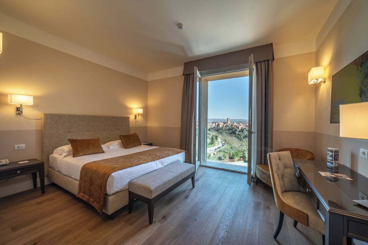 A hotel room with a large bed, a desk, and a chair, a view of a city through the window, and a ceiling light.