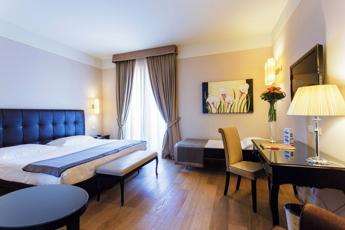 A hotel room with a large bed, a desk, a chair, a small table, and a desk lamp.