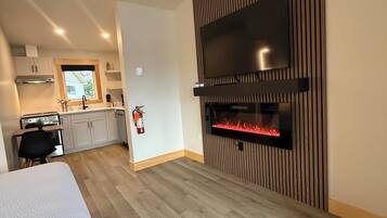 Una cocina moderna con estufa integrada, una zona de comedor con mesa y sillas, y una sala de estar con chimenea y un televisor grande montado en una pared de madera.