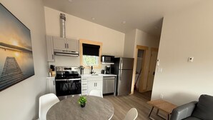 Cocina moderna con armarios blancos, estufa negra y microondas. Hay una mesa de comedor con sillas blancas y una planta pequeña. En la sala de estar se encuentra un sofá gris y una mesa de centro de madera.
