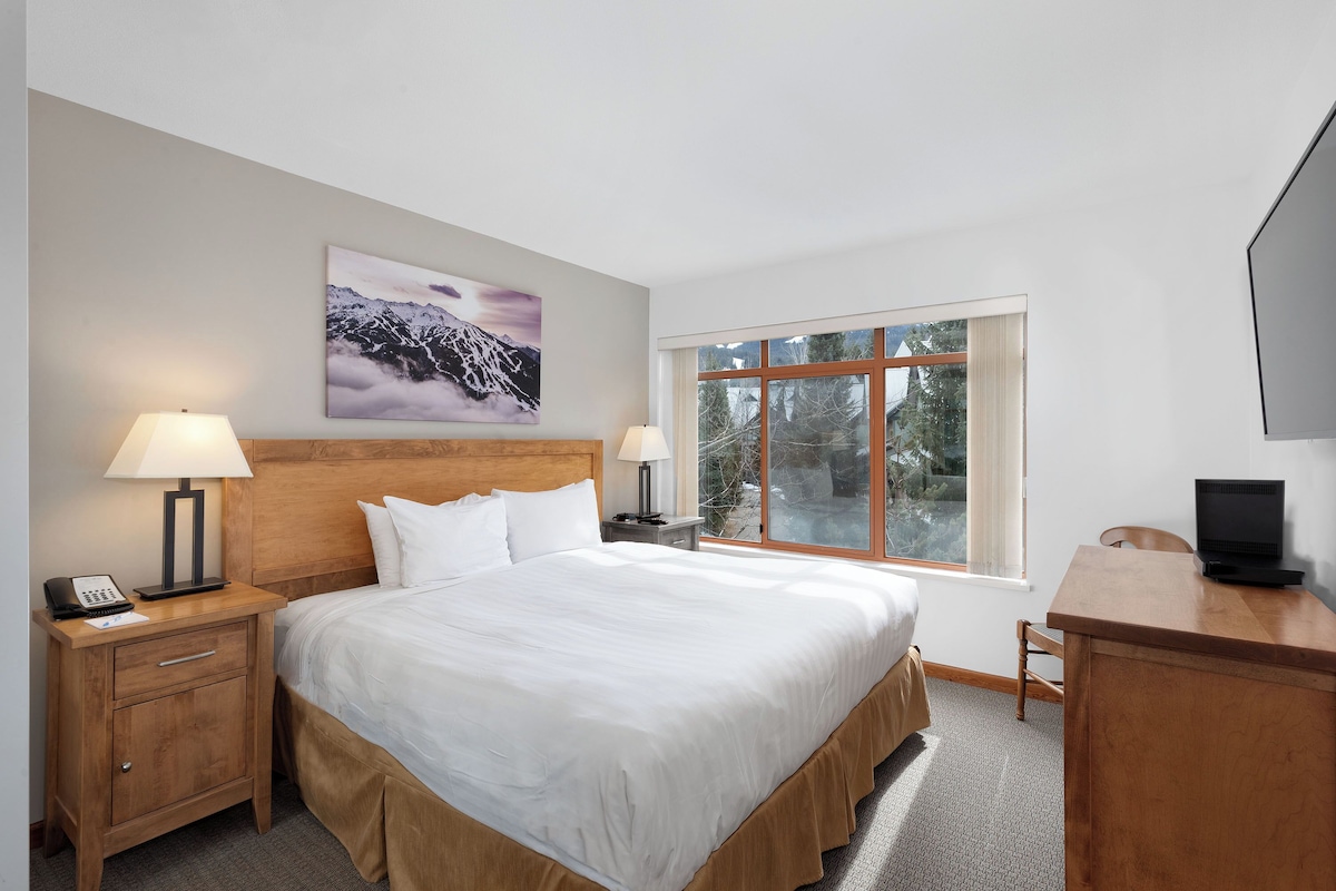 A hotel room with a large bed, two bedside tables with lamps, a desk with a laptop, a TV, and a window with a view of trees.