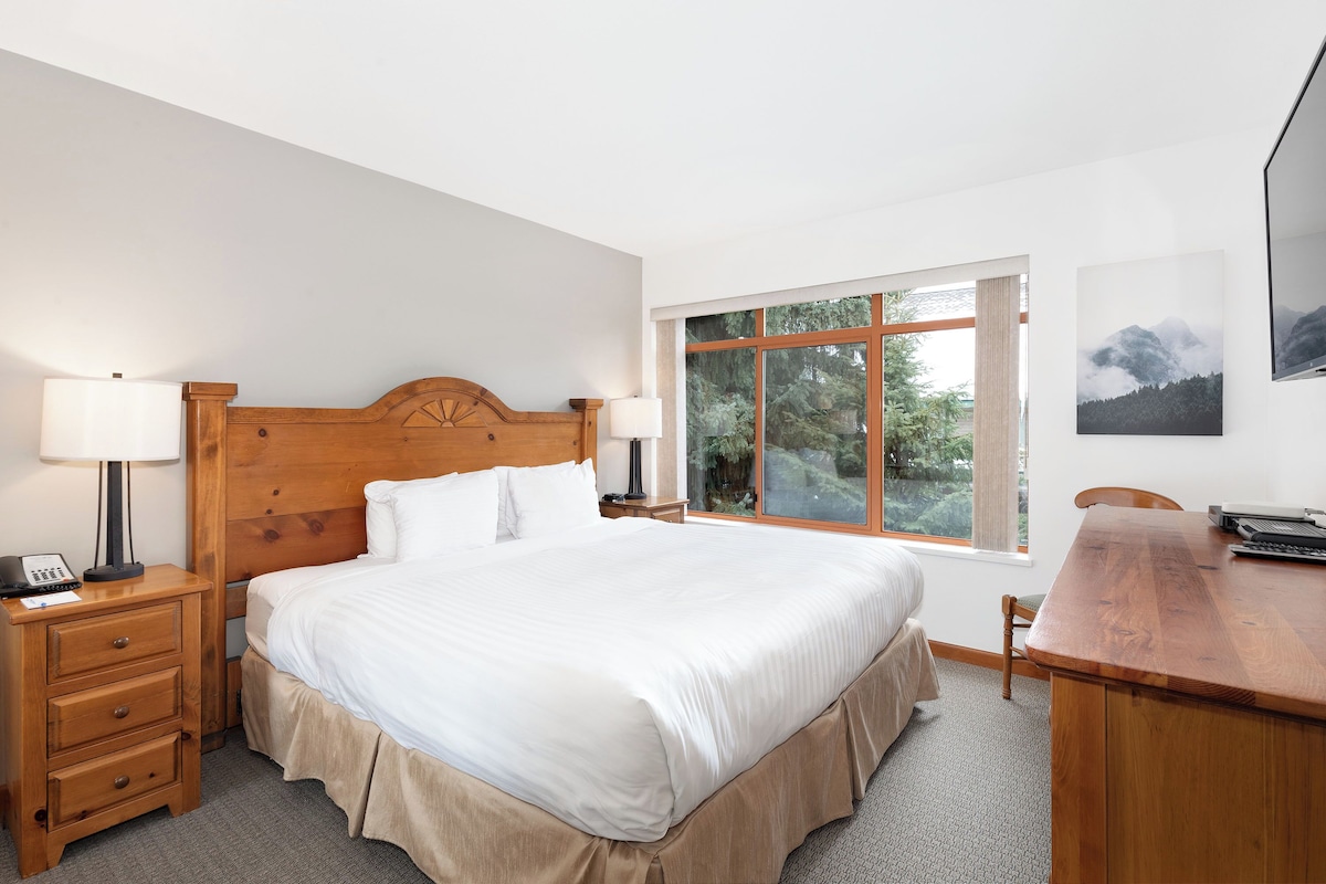A neatly arranged bedroom with a large bed, bedside tables with lamps, a wooden desk, and a window with a view of trees.