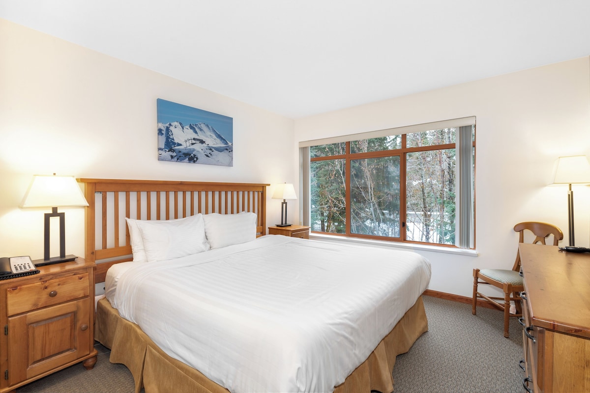 A neatly arranged bedroom with a large bed, wooden headboard, bedside tables with lamps, a window with a view of trees, and a painting on the wall.