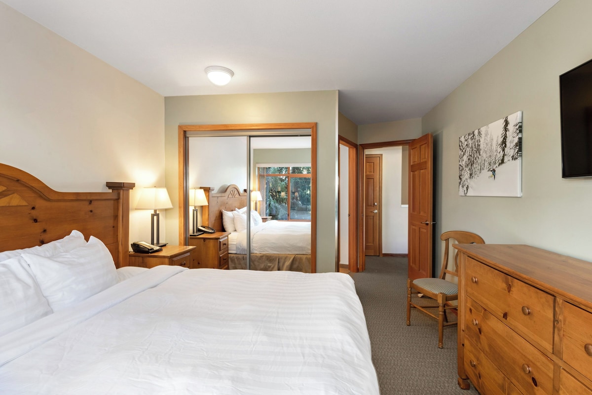 A bedroom with a large bed, wooden headboard, bedside tables with lamps, a dresser, and a mirror.