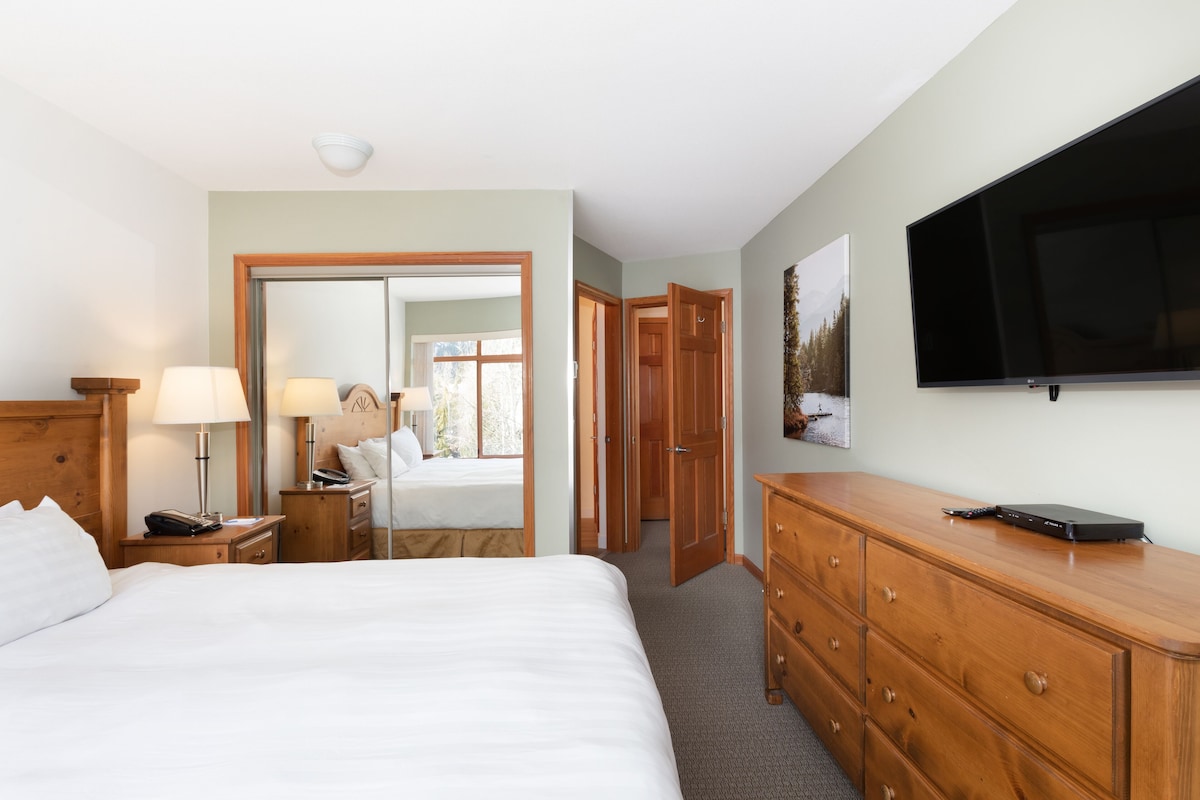 A hotel room with a bed, bedside tables, a dresser, a television, and a door leading to another room.