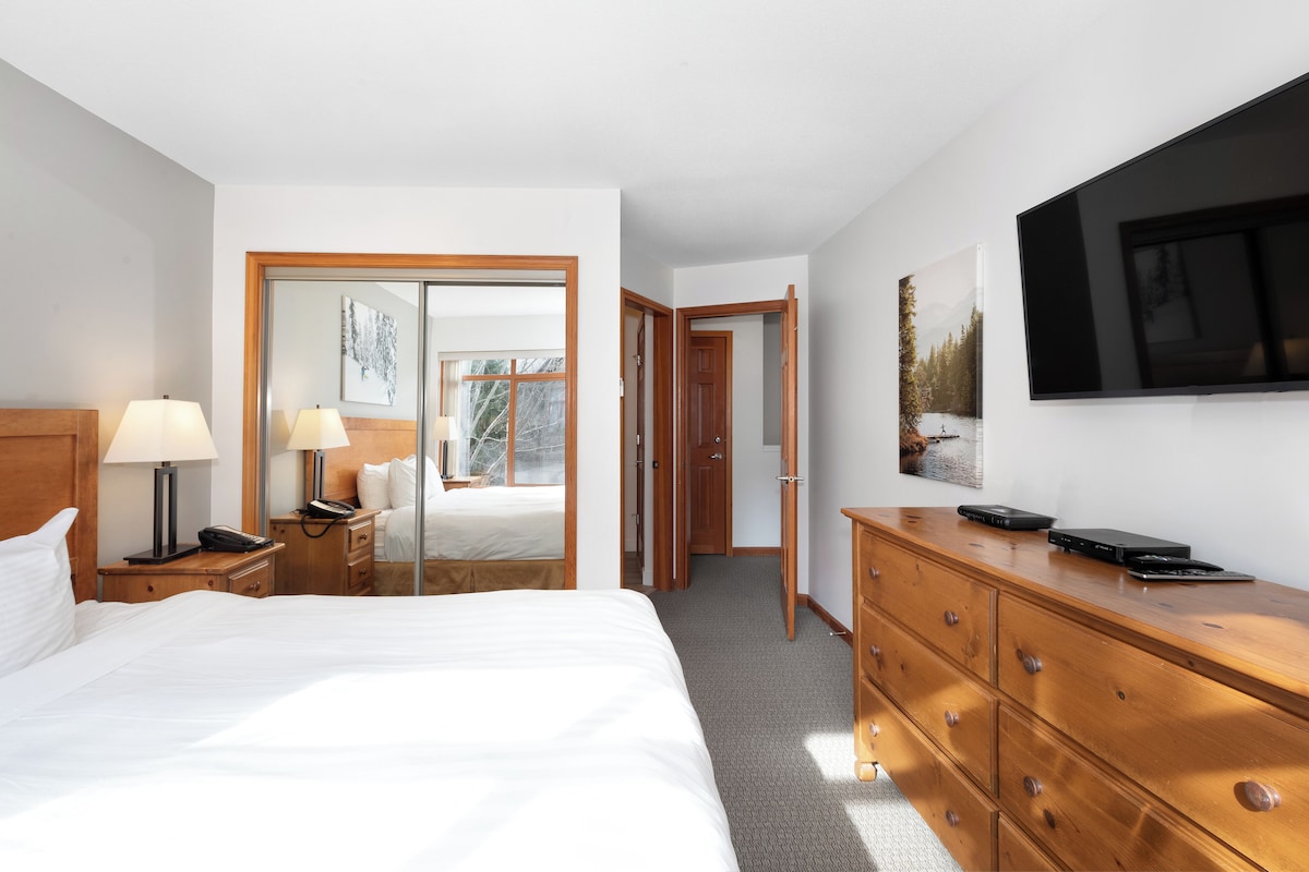 A hotel room with a large bed, a wooden dresser, a desk with a lamp, a telephone, and a flat-screen TV mounted on the wall.